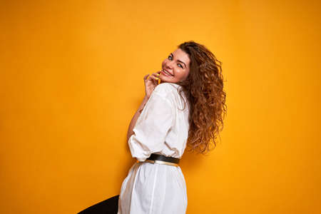 A beautiful girl on a yellow background smiles shyly. She standing sideways. She's wearing a white shirt. The hands are folded near the body. Smiling for the cameras.の写真素材