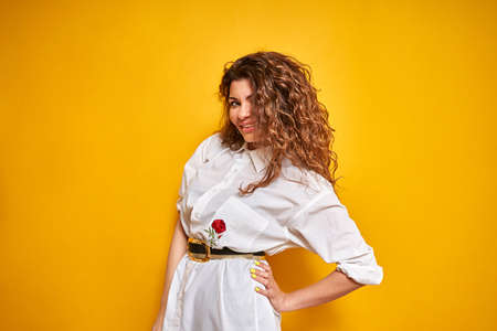 A young, sexy, confident girl with curly, lush hair in a white shirt poses on a yellow background. One hand rests on his side. Portrait of a young girl on a yellow background.の写真素材