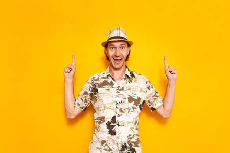 a young happy male tourist isolated on a yellow background points his index fingers up at an empty area. He is wearing a hat, a Hawaiian shirt. space for text. Concept - recreation, tourism, people.の写真素材