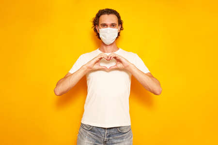 portrait of young man in medical mask folded his hands in form of heart in front of him on his chest. isolated on yellow background. space for text. concept of love, gratitude, sympathy, valentine dayの写真素材
