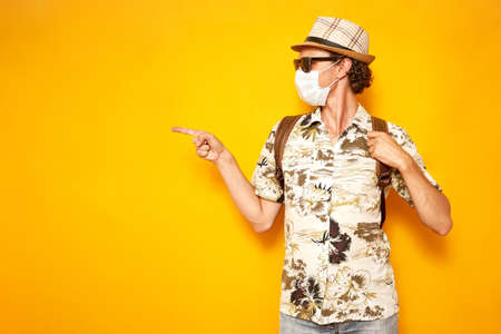 male tourist Hawaiian shirt, hat, sunglasses holds backpack with his hand and points his finger in direction of an empty space of text area to left yellow background. Traveling and covid19 vaccinationの写真素材