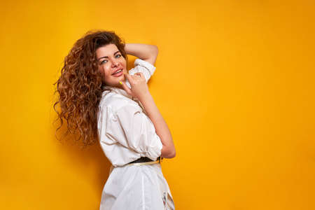 young curly haired woman in white shirt holds her hand to chin and adjusts her hair with thoughtful face stands sideways isolated on yellow background with place for text thinking about right decisionの写真素材