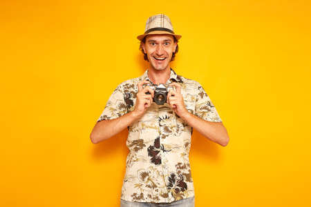 an excited male tourist traveler on vacation in hat and Hawaiian shirt holds camera in front of him and happy to see the resulting frame. isolated on yellow background with space for text. concept people. High quality photoの写真素材