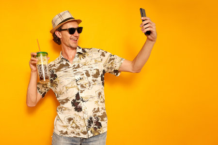 male tourist traveler in hat, Hawaiian shirt, sunglasses on vacation with drink in cocktail glass takes selfie on phone, communicates via video link. isolated on yellow background with space for text. High quality photoの写真素材