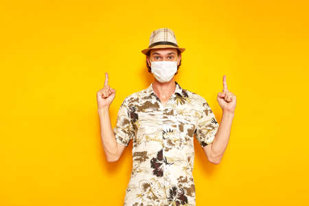 male tourist in medical mask points with his hands up at an empty advertising area above him. isolated yellow background. copy space. concept of people, travel, vacation, protection from covid19 virusの写真素材