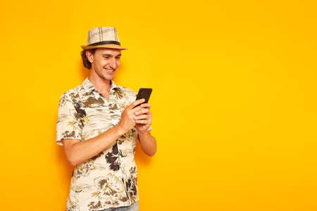 a male tourist on vacation with a smartphone in his hands writes a message and smiles. isolated on a yellow background. space for text. concept of people, recreation, communication in social networksの写真素材
