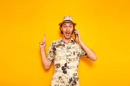 surprised, happy male tourist talking on smartphone points his finger up at an empty area for advertising. isolated on yellow background with space for text. concept of people, vacation, communicationの写真素材