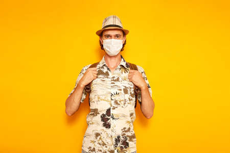 male tourist traveler with backpack in medical mask, Hawaiian shirt, hat holds straps with his hands and looks in front of him. isolated on yellow background. space for text. concept of people, travelの写真素材