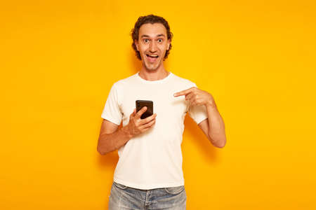 an excited happy man in casual clothes with phone in his hand points his finger at smartphone and we are happy to receive message about discounts, promotions and sales. isolated on yellow background.の写真素材