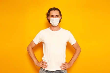 curly-haired young man in casual clothes wearing protective medical mask stands with his hands on his belt isolated yellow background with space for text. concept people, protection from virus, healthの写真素材