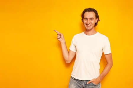 young curly haired smiling man in casual clothes points with his index finger at an empty area for advertising, promotions, discounts. isolated on yellow background with space for text. concept peopleの写真素材