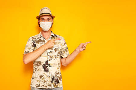 surprised male tourist traveler in medical mask points with his index fingers to side at an empty area for advertising. isolated on yellow background. copy space. concept people, vacation, advertisingの写真素材