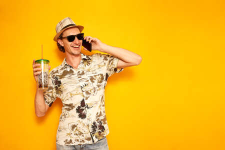 satisfied happy male tourist traveler with tropical drink in hand is talking on the phone and enjoying vacation abroad. isolated yellow background with space for text. concept recreation communicationの写真素材