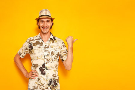 a happy smiling male tourist traveler on vacation points his thumb at an empty area for advertising. isolated on a yellow background with space for text. concept - people, adventures, flights abroadの写真素材