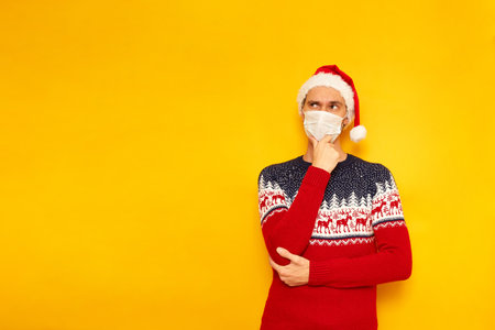 man in New Year sweater with reindeer, medical mask, Santa Claus hat holds his hand under his chin in thoughtful pose thinking about choice gifts. isolated yellow background with space for textの写真素材