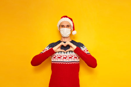 an excited man dressed in Christmas sweater with reindeer, red Santa Claus hat, medical mask folded his hands in shape of heart on his chest, sign love. isolated yellow background with space for textの写真素材