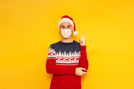 shocked, excited man in Christmas sweater with reindeer, medical mask, New Year Santa Claus hat, shows his index finger up, cool idea has come. isolated yellow background with space for text. holidayの写真素材
