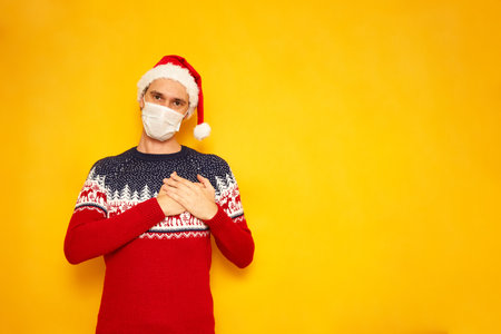 an excited man dressed in Christmas sweater with reindeer, red Santa Claus hat, medical mask folded his arms his chest on his heart, sign love, feelings. isolated yellow background with space for textの写真素材