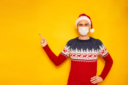 man in Christmas sweater, medical mask, red Santa Claus hat points with his index finger at an empty area for New Year advertising. isolated yellow background with copy space concept - people, holidayの写真素材
