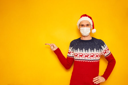 man in Christmas sweater, medical mask, red Santa Claus hat points with his index finger at an empty area for New Year advertising. isolated yellow background with copy space concept - people, holidayの写真素材