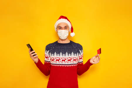 Online shopping, winter holidays, buy gifts concept. man thinking and holding credit card with mobile phone, wearing red Santa hat xmas sweater, medical mask, isolated yellow background space for textの写真素材