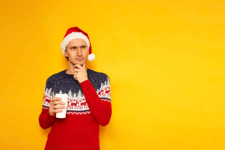 joyful man with paper cup for hot drinks coffee tea in New Year sweater Santa Claus hat points his finger empty advertising space isolated yellow background with space for text concept - good morningの写真素材