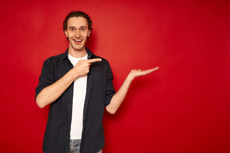 young, handsome, excited man in casual clothes, blue shirt, white T-shirt, points index finger towards hand free empty space for advertising text object isolated red background copy space. sale, promoの写真素材