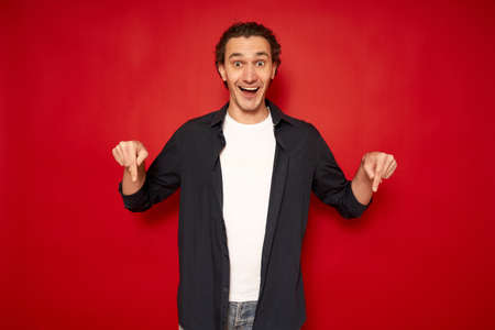 smile joyful guy pointing index fingers down advertising something standing posing in studio over red background space for text, smiling to camera. advertisement banner, look there concept. panorama.の写真素材