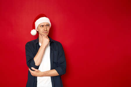 serious man in thoughtful pose holds his hand under his chin in Christmas Santa Claus hat isolated on red background with space for text concept - brainstorming, gift selection, holiday ideas, holidayの写真素材
