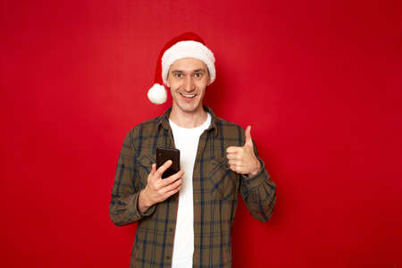 smiling man in casual clothes in Christmas hat with mobile phone in his hand shows thumbs up, approval sign, ok, app recommendation isolated on red background space for text concept, discounts, saleの写真素材