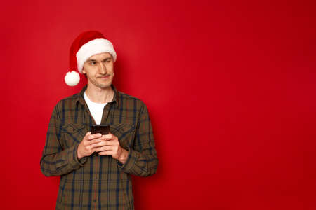 Satisfied hipster man with christmas hat types text message on cell phone, enjoys online communication, types feedback wears green shirt isolated on red studio wall. Technology, social digital conceptの写真素材