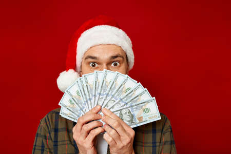 large studio portrait of young man in Christmas hat with wad of money in his hands holding fan of dollars in front of his face in shock from successful lottery win. concept winning, cash back, wealthの写真素材