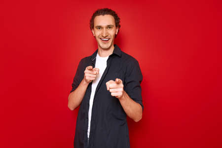 smiling, excited man points his index fingers forward in front of him. he is dressed in casual clothes, blue shirt, white T-shirt. isolated on red studio background. concept - people, victory, bestの写真素材