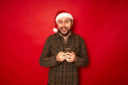 a happy excited man in a Santa Claus Christmas hat with a phone in his hands rejoices at received sensation. isolated on red studio background. concept - new year, application, internet, communicationの写真素材