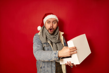 surprised excited man in Christmas hat, denim jacket opens white box holding it in his hands shocked by gift received for new year. isolated red studio background. concept - holidays, emotions, peopleの写真素材