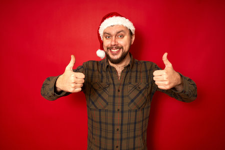 smiling man in Christmas hat, casual clothes shows thumbs up with both hands isolated on red studio background with space for text. concept - holidays, new year, promotion, discounts, favorable offerの写真素材