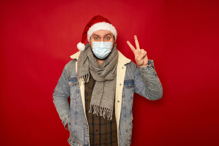 man in Christmas hat, medical mask on face shows peace victory sign hand isolated studio red background space for text concept - holidays, medicine, health, protection from infections, coronavirusの写真素材