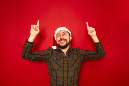 portrait of smiling man in Christmas hat casual clothes pointing his index fingers up at an empty space for advertising text isolated on red studio background concept people, recommendation, gesturesの写真素材