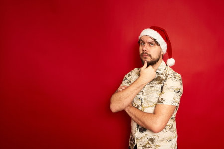 Puzzled confused young traveler tourist man in summer casual clothes christmas santa hat put hand on chin isolated on red studio background. Passenger traveling on holiday. Air flight journey conceptの写真素材