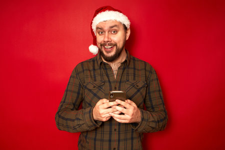 Wow, I got message from stranger. Fashionable guy in santa hat and green shirt, holds cell phone, gasps from surprisement, reads stunning news, poses over red wall with empty space for informationの写真素材
