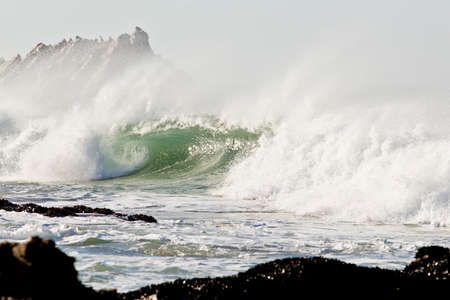 Strong wind blowing sea spray off a big wave with rocks behind and in front
