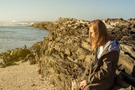 Woman leaning against rocks at the beach on a cool winters dayの写真素材