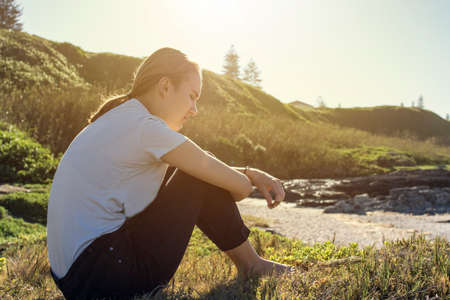 young woman sitting and relaxing in soft morning sun on wild grass next to the beach at low tideの写真素材