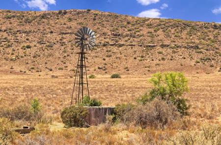 Old broken overgrown windmill standing unused with thorn trees and bushes growing out of the reservoir in the Karoo landscapeの写真素材