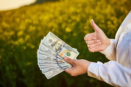 Agronomist woman or farmer holding money dollar banknote with blossoming cultivated canola field in...