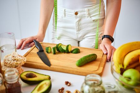Young happy woman cutting cucumber for making salad in the beautiful kitchen with green fresh ingredients indoors. Healthy food and Dieting concept. Loosing Weightの写真素材