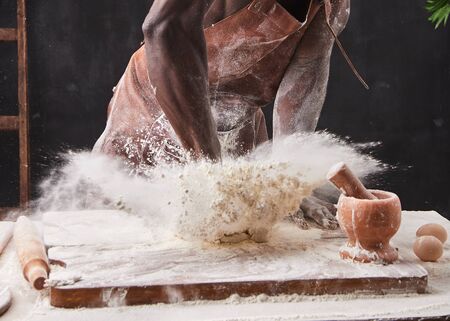 Dark skin baker man kneads Dough in the kitchen. White flour flies in air on black background,...