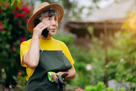 Senior woman gardener in a hat working in her yard and speaks on the phone. The concept of gardening, growing and caring for flowers and plants.の写真素材