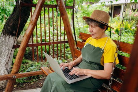 Senior woman gardener in a hat is watching something on a laptop computer and making a video call in the yard while outdoors.の写真素材