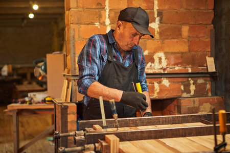 Portrait of a senior carpenter in uniform gluing wooden bars with hand pressures at the carpentry...